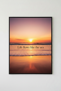 Framed artwork with a sunset over water and the text 'Life flows like the sea' on a white wall.